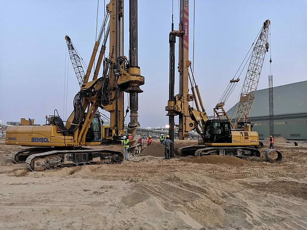 Two rotary piling rigs working side by side on an industrial site at dusk